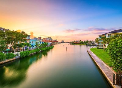 Scenic view of a calm canal flanked by lush greenery and colorful homes under a vibrant sunset sky.