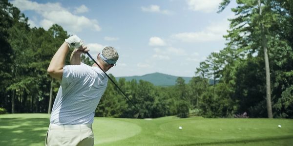 Man swinging golf club on a sunny golf course with trees and mountains.