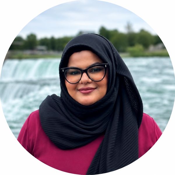 Smiling woman wearing glasses and a black hijab near a waterfall.