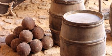 Stack of cannonballs and two wooden barrels on a dirt ground.