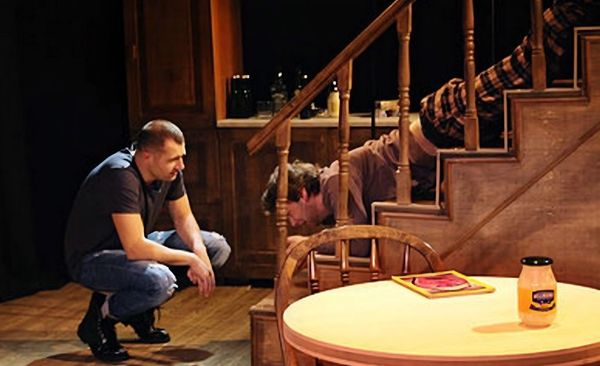Two men interacting near a staircase in a dimly lit room.