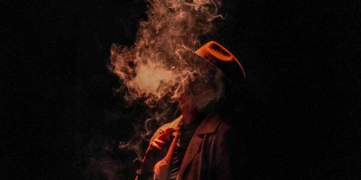 Mysterious figure in a hat enveloped by swirling smoke in dim light.