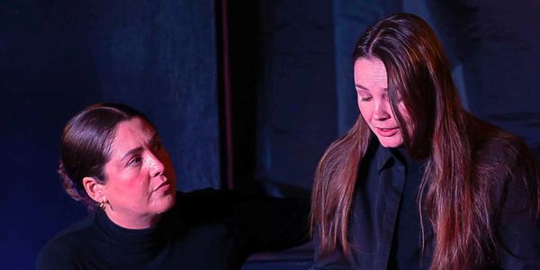 Two women in black engaged in a serious conversation in a dimly lit setting.
