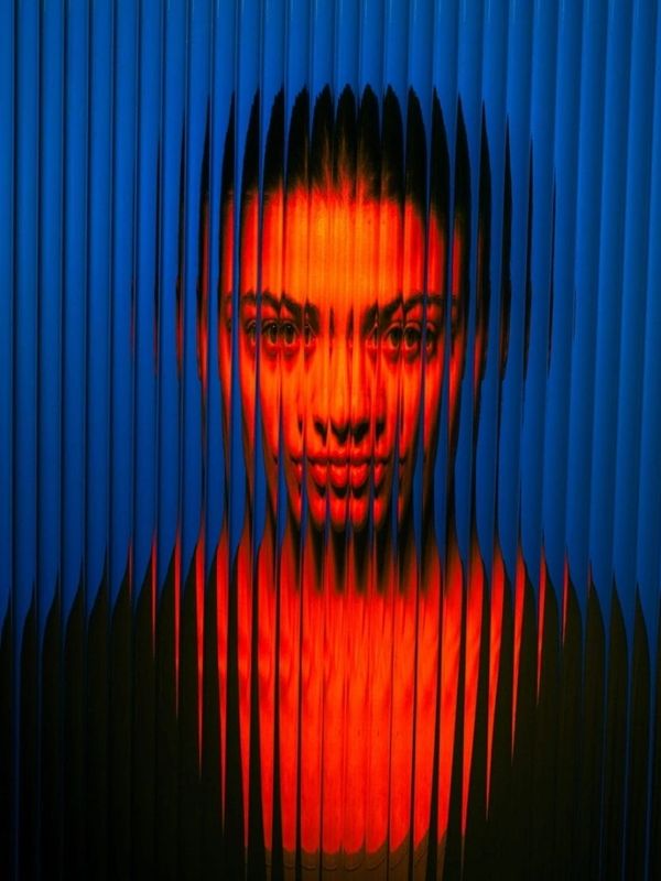 A woman's face distorted behind vertical slats with red and blue lighting.