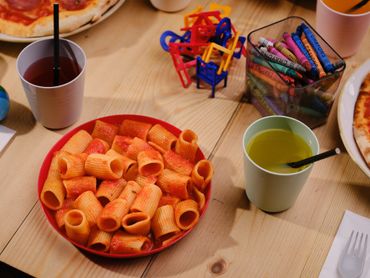 A plate of pasta with two drinks and crayons on a wooden table.