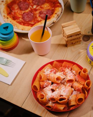 Plate of rigatoni pasta with tomato sauce and grated cheese, pizza, and a drink on a wooden table.