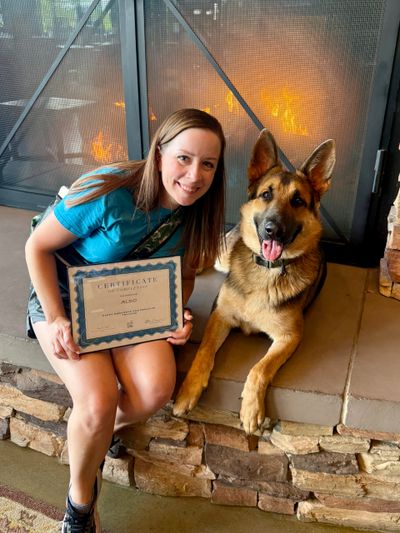 Woman proudly holds a certificate sitting next to a German Shepherd by a fireplace.
