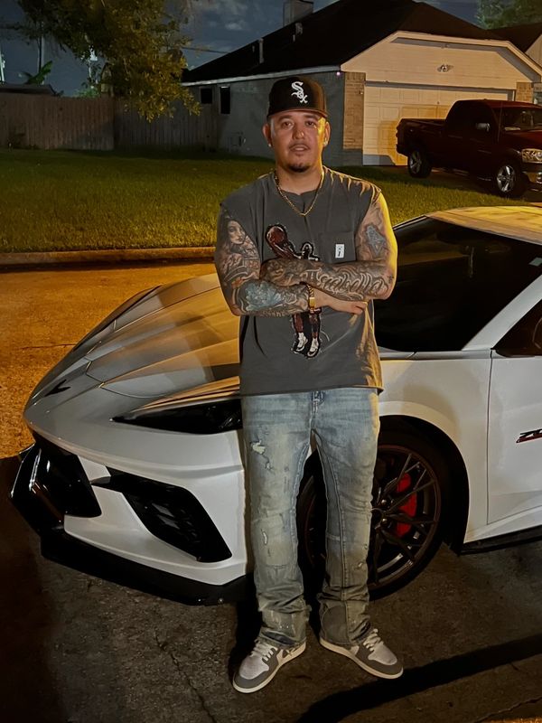 Man with tattoos stands confidently in front of a white sports car at night.