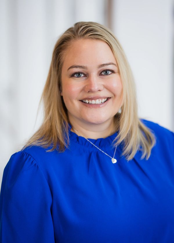 Smiling woman with blonde hair in a bright blue blouse and heart necklace.