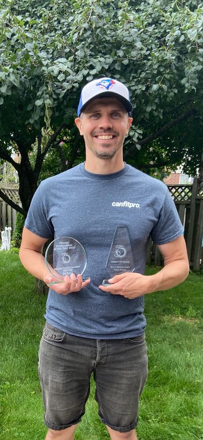 Man smiling, holding two fitness awards outdoors.