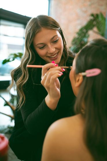 A woman happily applies makeup to another person indoors.