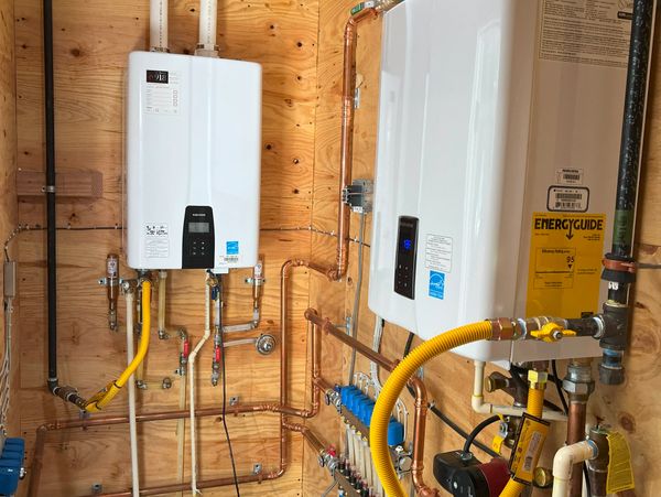 Two white Navien boilers with copper and PVC piping on a wooden wall.