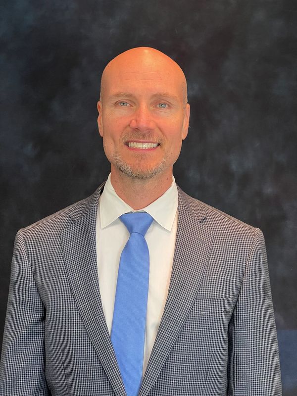 Smiling bald man in checkered suit with blue tie against dark background.