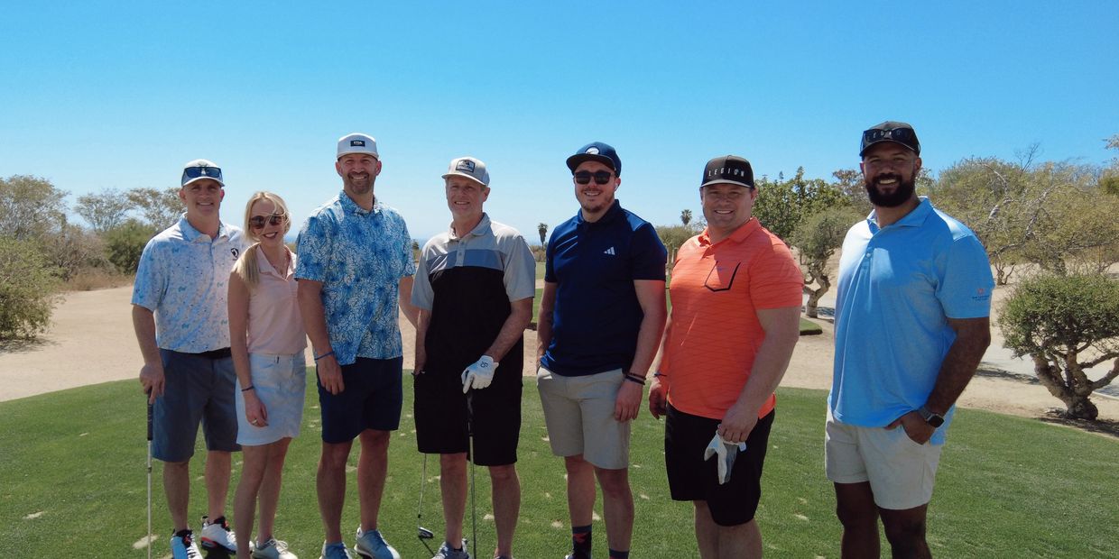 Group of seven people posing on a golf course under clear blue sky.