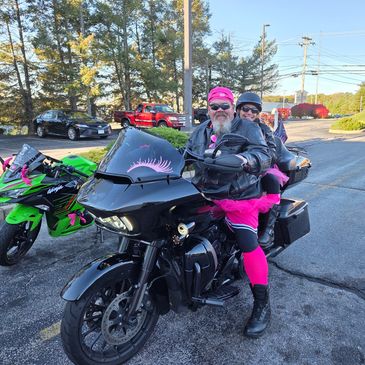 Two people on a black motorcycle dressed in pink accessories.