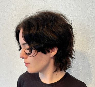 Profile of a person with medium-length wavy dark hair against a white wall.