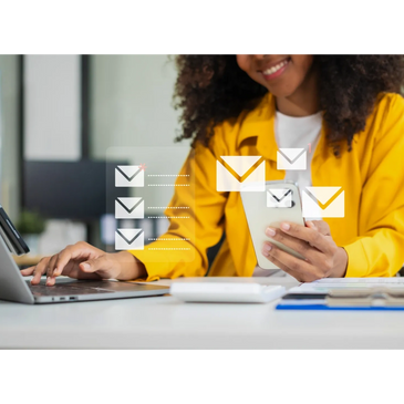 Woman in yellow jacket checking emails on phone with laptop nearby.