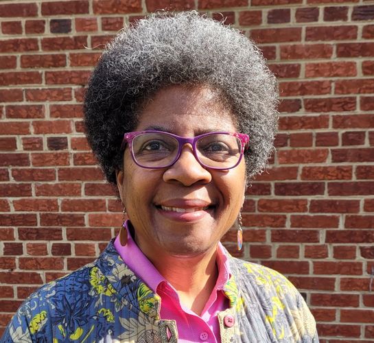 Smiling woman with curly gray hair and purple glasses standing against a brick wall.
