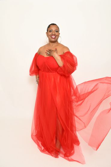 Woman in a flowing red off-shoulder gown smiling against a white background.