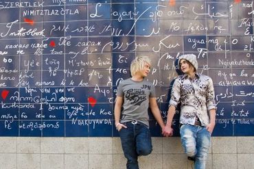 A couple holding hands in front of a wall with phrases in different languages.