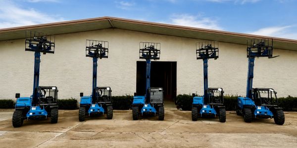 Five blue forklifts with raised forks in front of a warehouse.