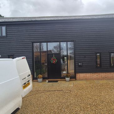 Modern house with black wood siding and glass entrance, flanked by vehicles on a gravel driveway.