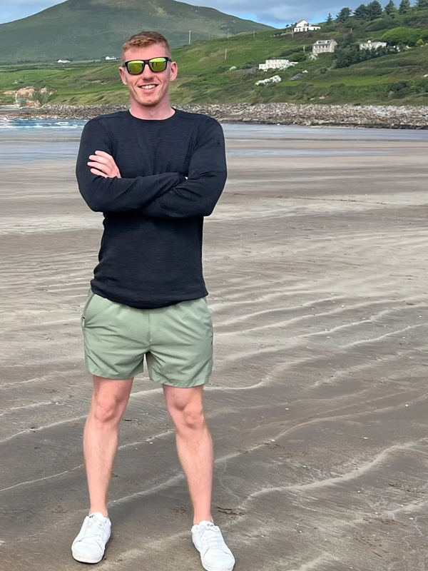 Man in sunglasses standing on a beach with hills in the background.