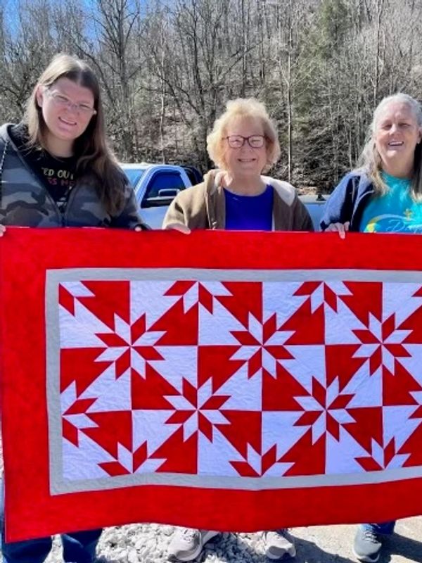Three women holding a red and white quilt outdoors.