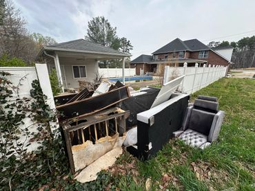 Discarded furniture and debris piled by a white fence near houses.