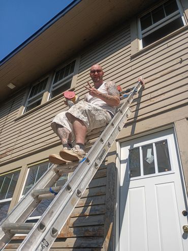 Man sitting on a ladder painting a house, making a peace sign.