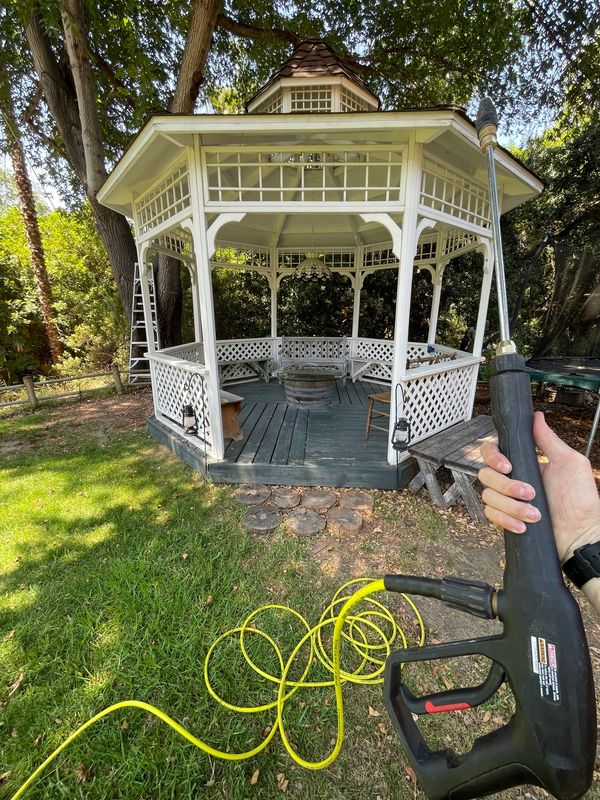 A white gazebo in a garden with a person holding a power washer.
