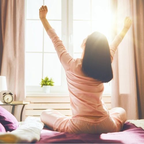 A picture of a person sitting on the side of the bed stretching their arms up toward the air while looking out a window.