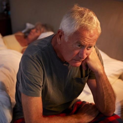 Man sitting on the side of the bed at night looking frustrate, while his partner sleeps behind him.