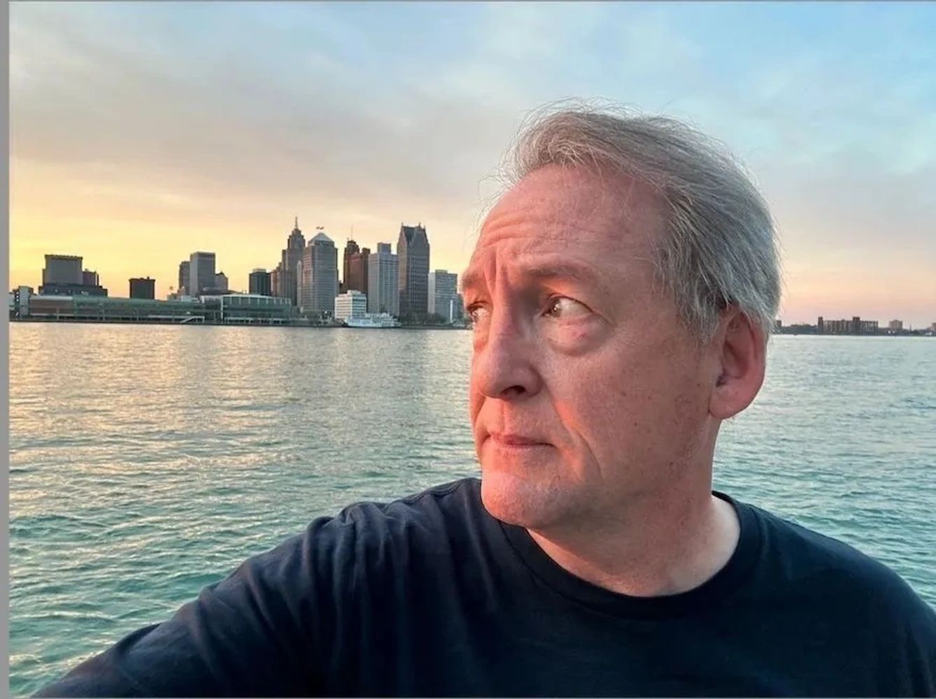 Man looking thoughtfully across the water at a city skyline during sunset.
