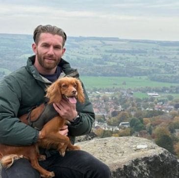 Man holding a happy dog with a scenic countryside view in the background.