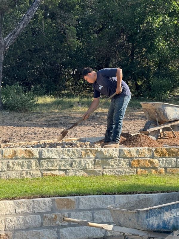 A man shovels gravel on a stone retaining wall in a sunny outdoor setting.