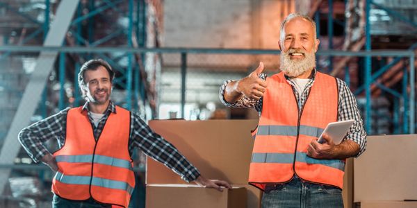 Two warehouse workers in orange safety vests smiling and giving thumbs up.