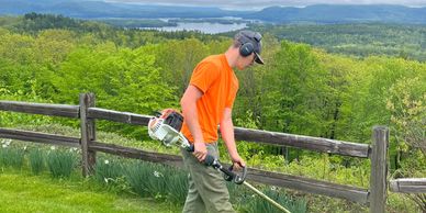 Crew member using a weed whacker to trim tall grass under fence.