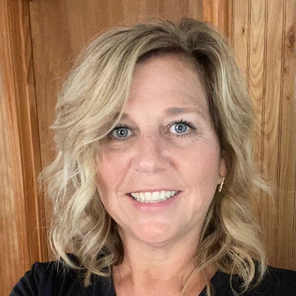 Smiling woman with blonde wavy hair wearing a black shirt in front of wooden panels.