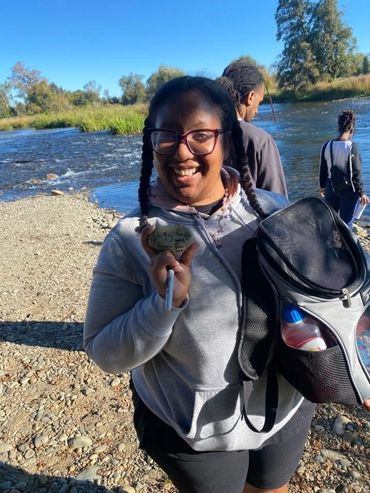 A smiling woman holding a rock by the river on a sunny day.