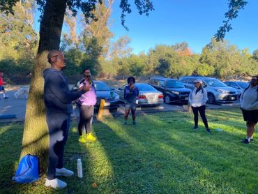 A group of people standing in a park, listening to a speaker near a tree.