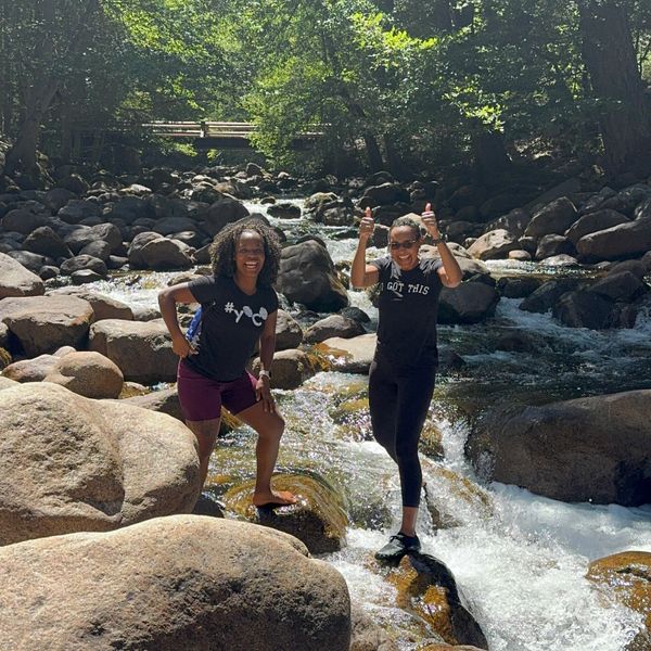 Two women joyfully pose on rocks by a flowing river in a sunny forest.