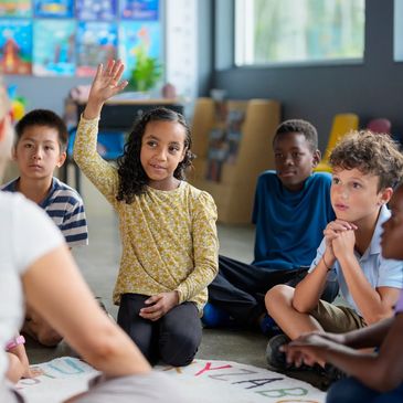 A young girl raises her hand in a classroom while other children listen attentively.