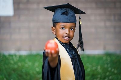 Kindergarten graduation. Young man holding apple and wearing graduation cap and gown.