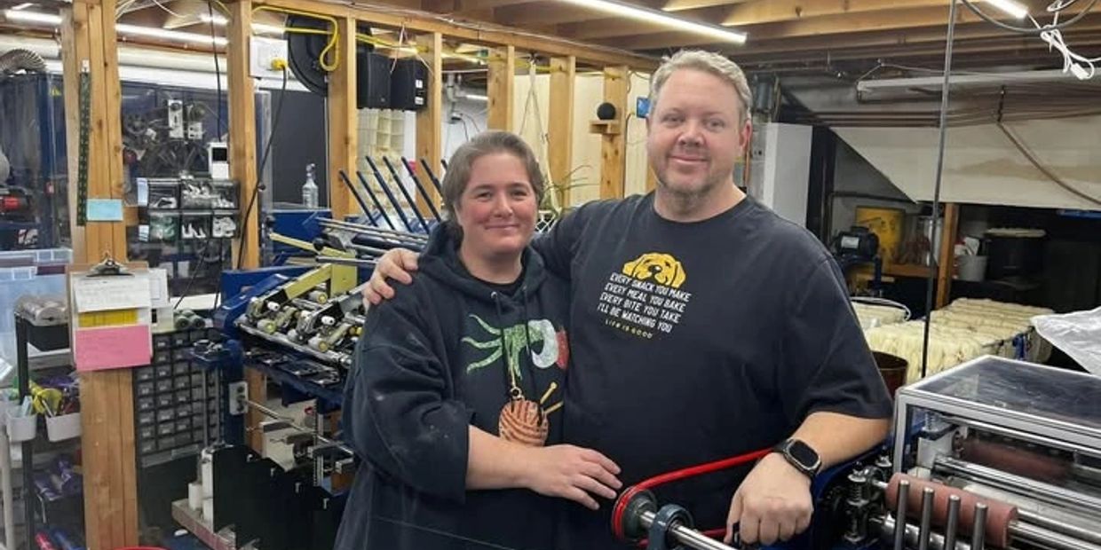 A couple stands together in a workshop filled with machinery and tools.
