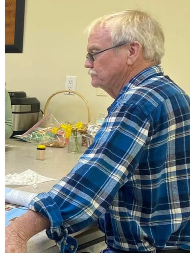 Older man in a blue plaid shirt standing at a counter in a casual setting.
