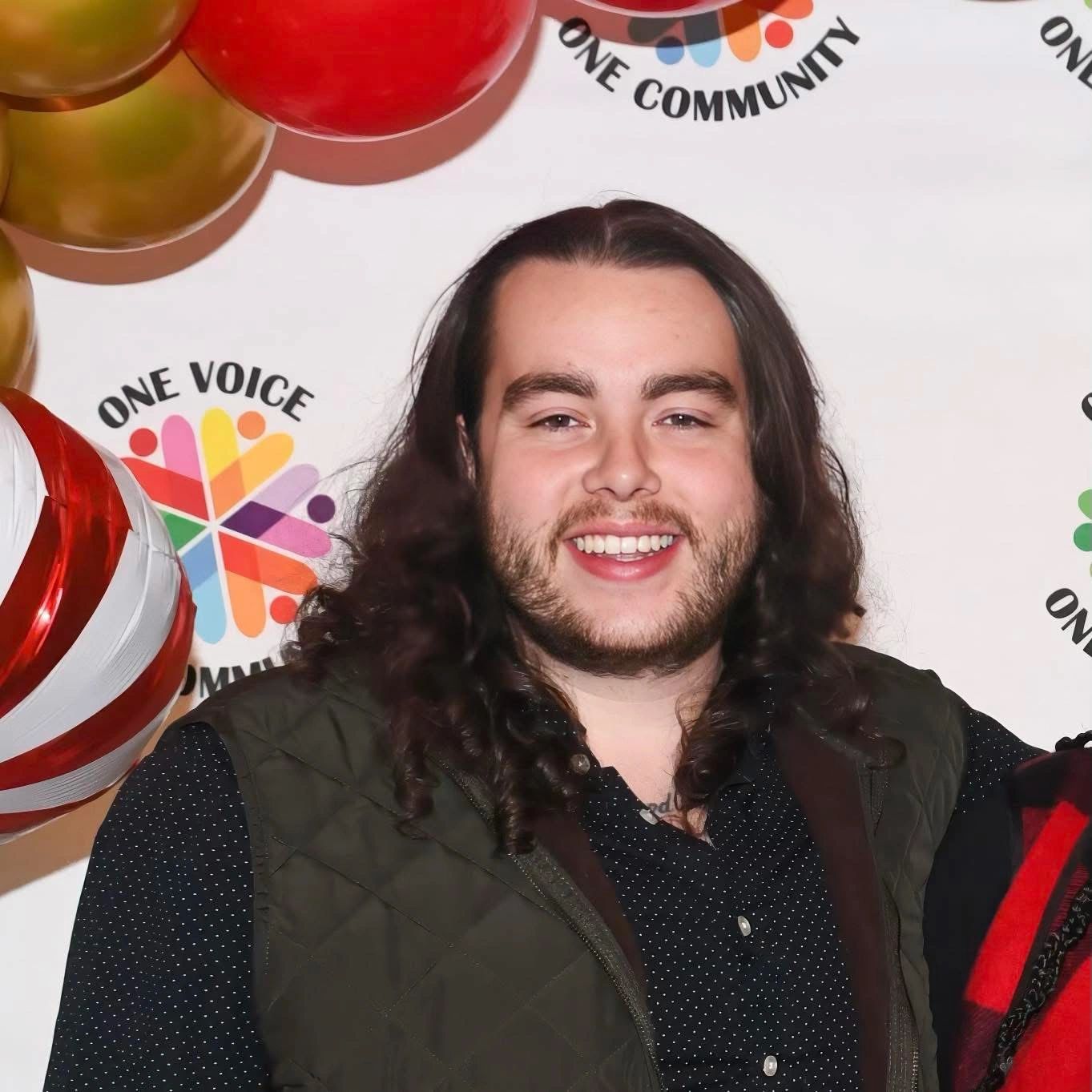 Smiling man with long hair at a festive event with colorful balloons.