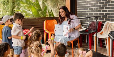 A woman reads a book to a group of young children in an outdoor setting.