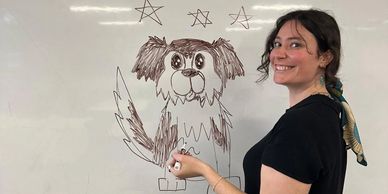 Woman smiling by a whiteboard drawing of a dog with stars above.