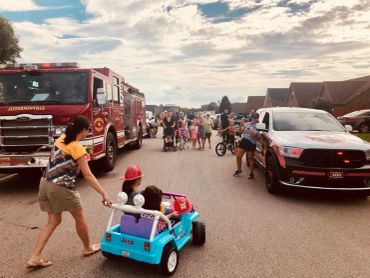 A woman pulls children in a toy car during a street parade with fire truck and crowd.